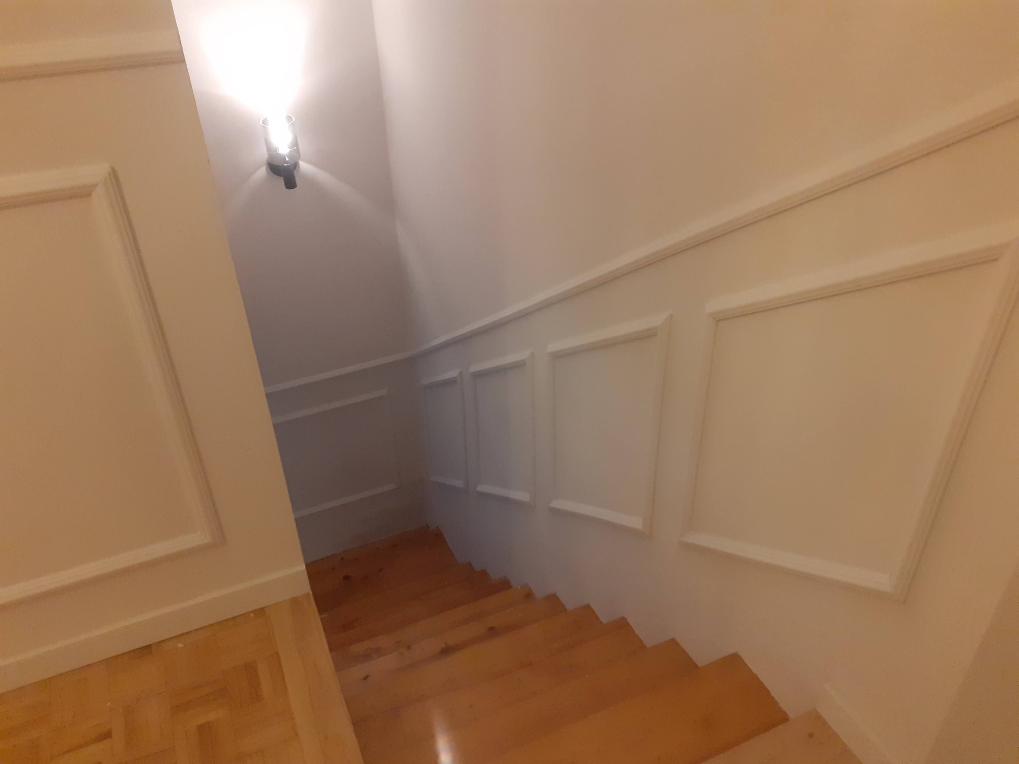 Klatka schodowa z drewnianymi schodami i ścianami z białymi panelami dekoracyjnymi oraz oświetleniem punktowym.