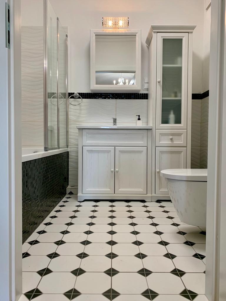 Biała łazienka z czarno-białą podłogą w karo, kabiną prysznicową z czarnymi płytkami mozaikowymi i białymi meblami łazienkowymi z lustrem.