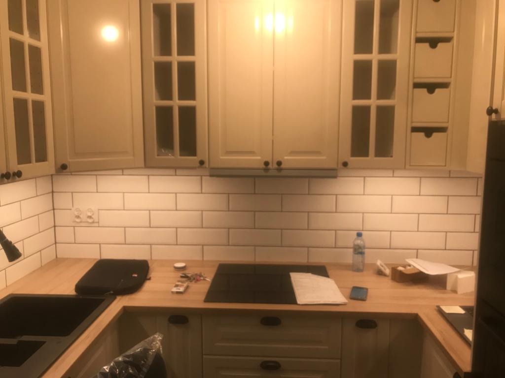 Jasna kuchnia z białymi płytkami metra, drewnianym blatem i czarnym zlewem, przedłużona lampa sufitowa, butelka wody i telefon na blacie.