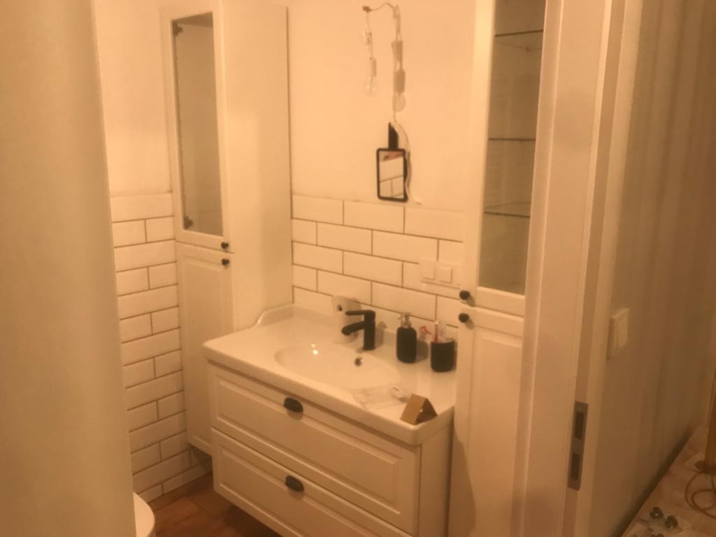 Biała łazienka z umywalką nablatową, szafką z szufladami, wysoką szafką z przeszklonym frontem i białymi płytkami na ścianie, oświetlenie w trakcie montażu.