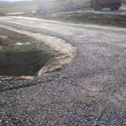 Nowo utwardzona droga z szarego kruszywa, widoczny wykop ziemny po lewej stronie z wystającą rurą odprowadzającą wodę, w tle sprzęt budowlany i ciężarówka, pagórkowaty krajobraz.