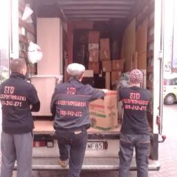 Trzech mężczyzn w odzieży roboczej z napisem 'STD Przeprowadzki' na plecach, stojących przed otwartym tyłem ciężarówki wypełnionej kartonami i meblami podczas załadunku.