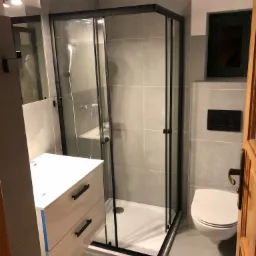 Szara łazienka po remoncie z kabiną prysznicową typu walk-in z czarnymi ramami, białą szafką umywalkową z czarnymi uchwytami i wiszącą toaletą.