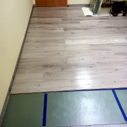 Podłoga z paneli w trakcie układania, widoczna mata podkładowa zabezpieczona taśmą, przy ścianie ułożone panele imitujące jasne drewno.