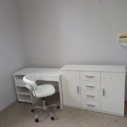 Białe biurko z szafką i krzesłem na kółkach w pustym, jasnym pokoju z płytkami podłogowymi.