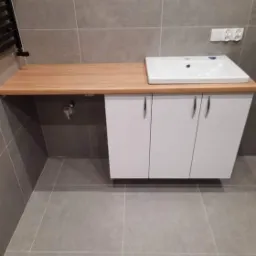 Łazienka z drewnianym blatem pod umywalką, białe szafki podwieszane, szare płytki na ścianach i podłodze, widoczny grzejnik drabinkowy i gniazdko elektryczne.