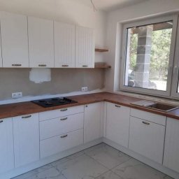 DamianMeble - Jasna kuchnia z białymi frontami szafek, drewnianym blatem i widokiem na zewnątrz przez okno. Ściana nad blatem nie jest jeszcze wykończona.