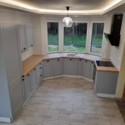 Szara zabudowa kuchenna z drewnianym blatem, widok na okno z zewnątrz, nowoczesny okap podszafkowy i ceramiczna podłoga imitująca drewno.