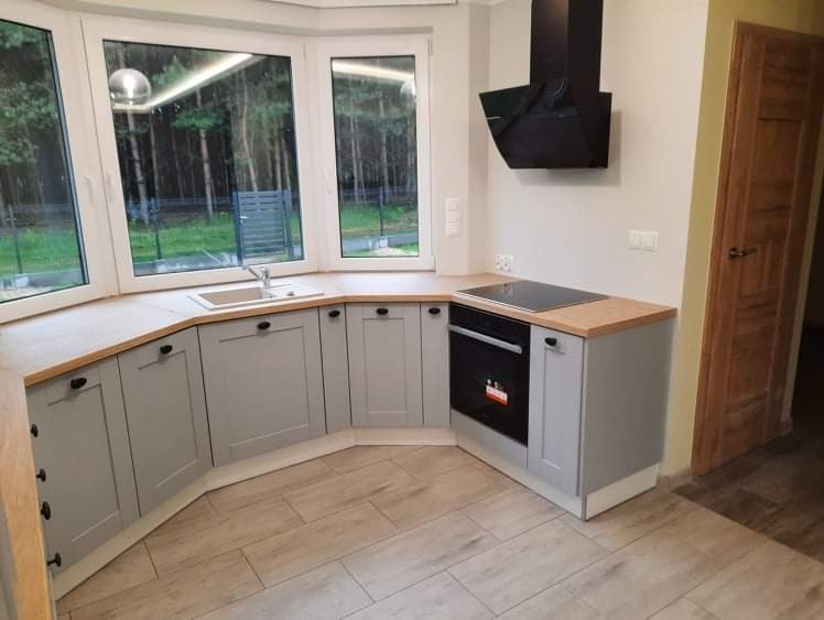 Jasnoszara kuchnia na wymiar z drewnianym blatem, widok na las przez okno wykuszowe, czarny okap i płyta indukcyjna.