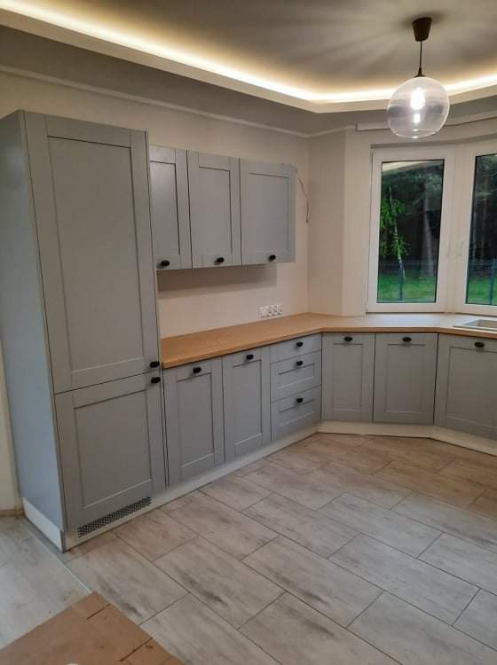 Szara zabudowa kuchenna z drewnianym blatem, czarne okrągłe uchwyty, widok na okno i szklaną lampę wiszącą, podłoga wyłożona panelami imitującymi drewno.