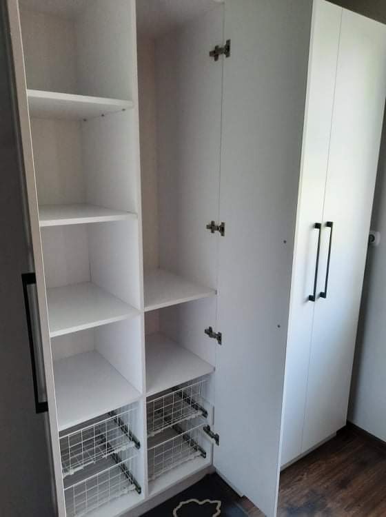 Biała szafa z otwartymi drzwiami, widoczne półki i wysuwane metalowe kosze, czarne uchwyty, minimalistyczny design.