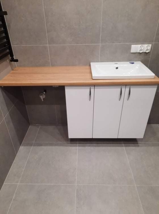 Łazienka z drewnianym blatem pod umywalką, białe szafki podwieszane, szare płytki na ścianach i podłodze, widoczny grzejnik drabinkowy i gniazdko elektryczne.