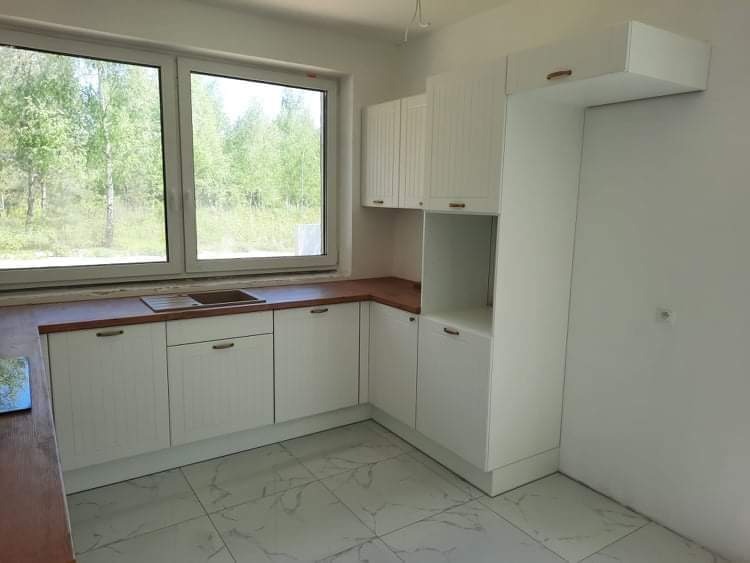 Jasna kuchnia z białymi, frezowanymi frontami szafek i drewnianym blatem, widok na zielony las za oknem.