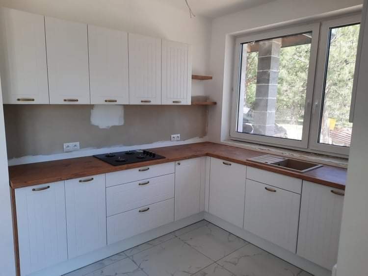 Jasna kuchnia z białymi frontami szafek, drewnianym blatem i widokiem na zewnątrz przez okno. Ściana nad blatem nie jest jeszcze wykończona.