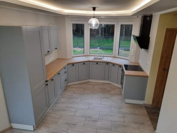 Szara zabudowa kuchenna z drewnianym blatem, widok na okno z zewnątrz, nowoczesny okap podszafkowy i ceramiczna podłoga imitująca drewno.