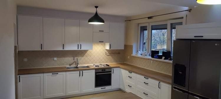 Jasna kuchnia z białymi szafkami z czarnymi uchwytami i drewnianym blatem, oświetlona wiszącą, czarną lampą