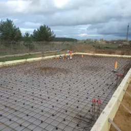 Zbrojenie płyty fundamentowej z widocznymi pionowymi prętami, deskowaniem z izolacją termiczną i instalacją kanalizacyjną, na tle ogrodzenia i pochmurnego nieba.