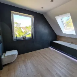 Wykończona łazienka na poddaszu w Pogwizdowie z czarnymi płytkami na ścianach, jasną podłogą imitującą drewno, podwieszaną toaletą i wanną zabudowaną w czerni z oświetleniem LED. Widok na zewnątrz...