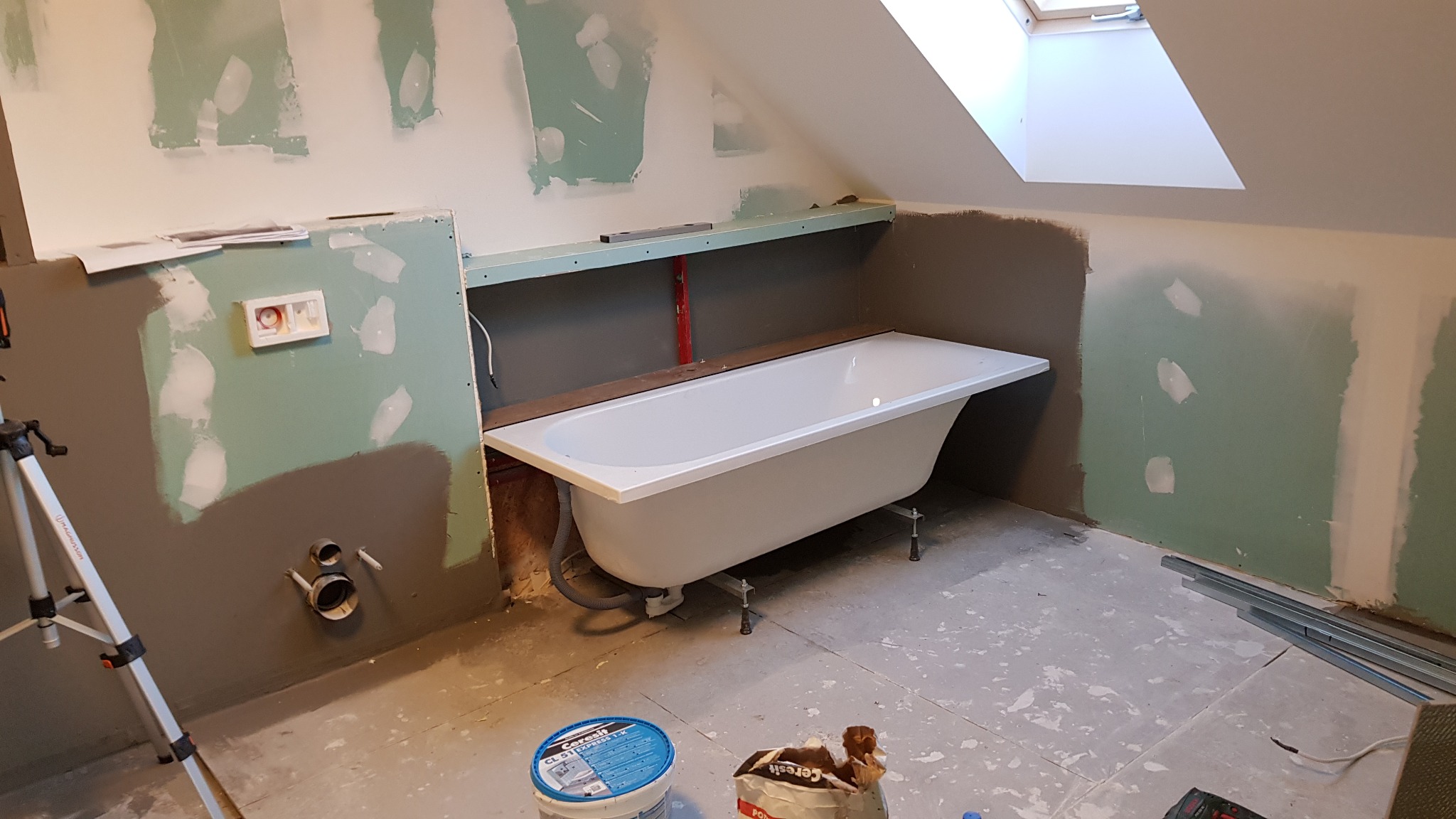 Biała wanna na nóżkach w trakcie remontu łazienki na poddaszu, widoczne płyty karton-gips, niezaprawione ściany, rury kanalizacyjne, poziomica i materiały budowlane.