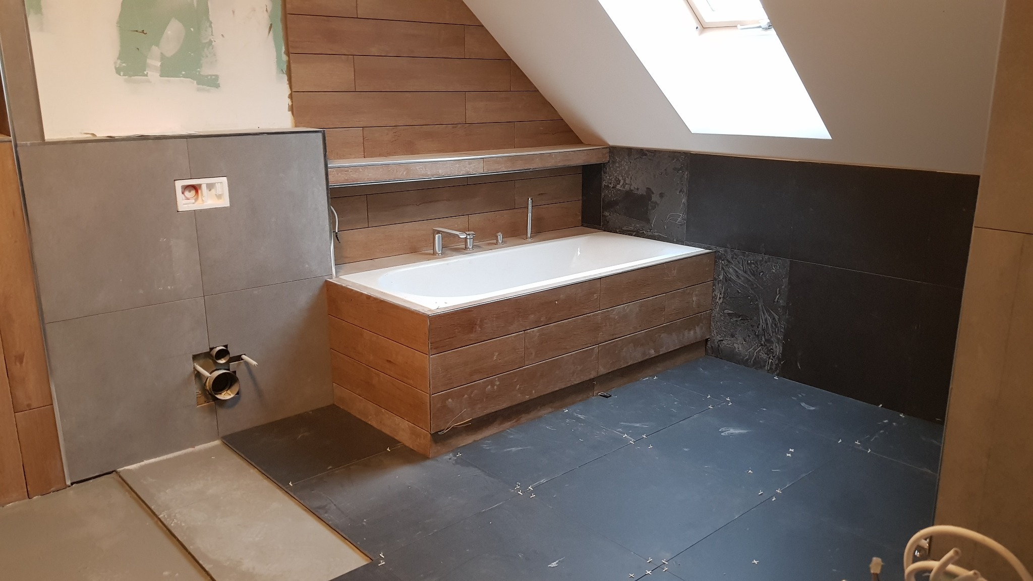 Łazienka w trakcie remontu: wanna obudowana drewnem, częściowo ułożone ciemne płytki podłogowe i szare płytki ścienne w strefie umywalki, widoczne instalacje wodno-kanalizacyjne, okno dachowe...