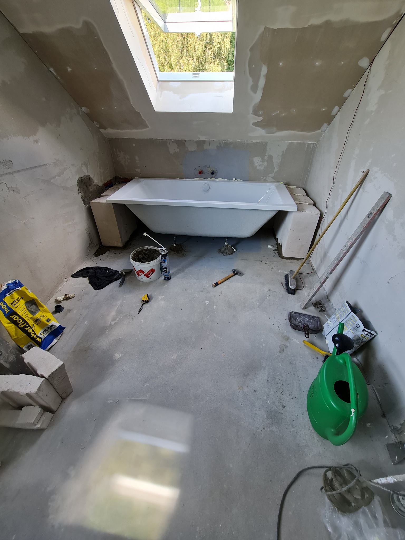 Biała wanna osadzona na bloczkach w surowej łazience ze skośnym sufitem i oknem dachowym, widoczne narzędzia i materiały budowlane.