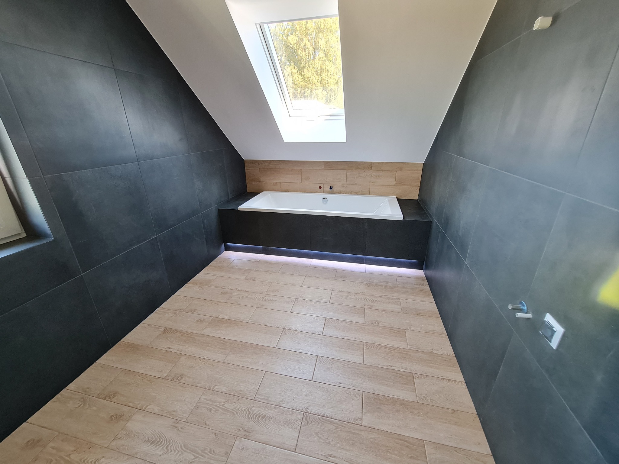 Nowoczesna łazienka na poddaszu z oknem dachowym, wykończona ciemnymi płytkami na ścianach i jasnymi panelami podłogowymi imitującymi drewno, z białą wanną podświetloną od dołu.