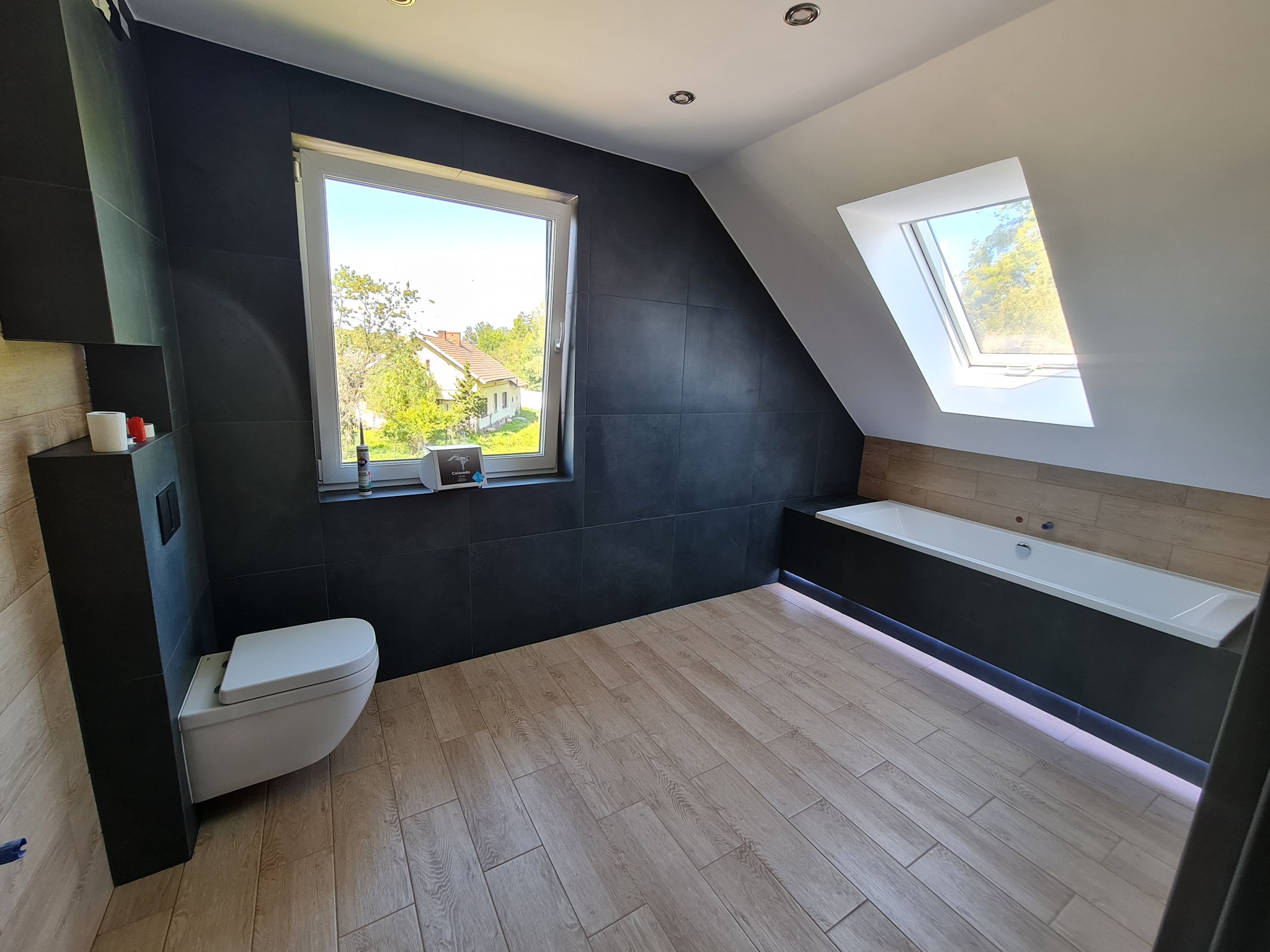 Wykończona łazienka na poddaszu w Pogwizdowie z czarnymi płytkami na ścianach, jasną podłogą imitującą drewno, podwieszaną toaletą i wanną zabudowaną w czerni z oświetleniem LED. Widok na zewnątrz...