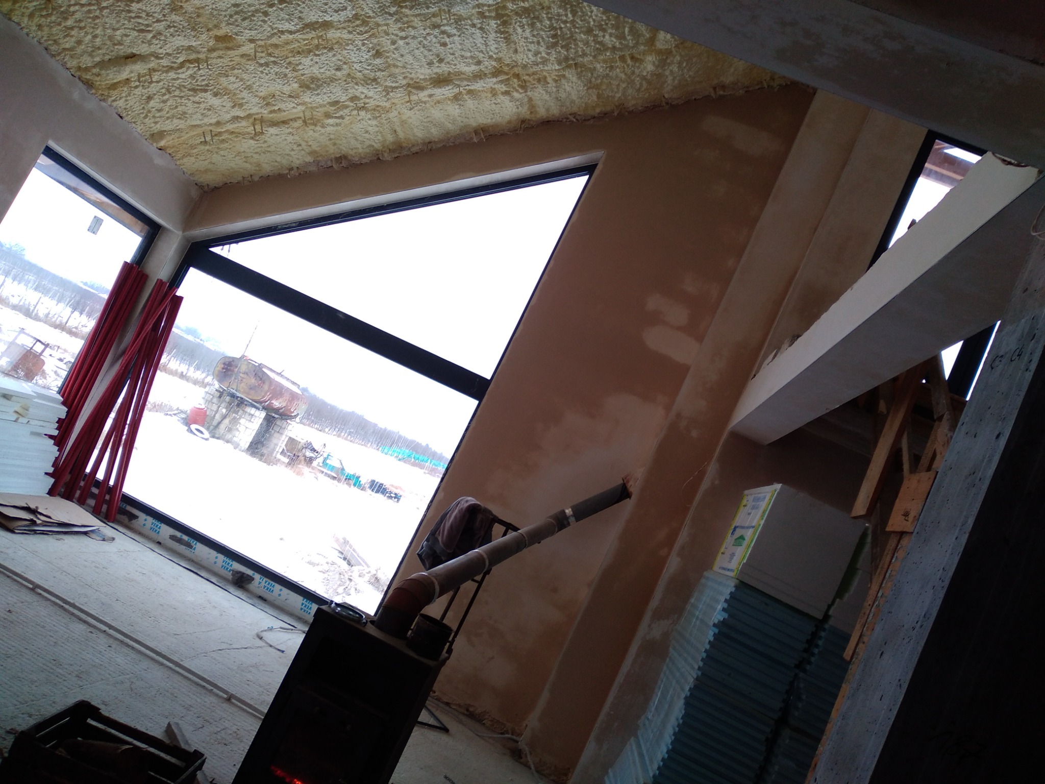 Wnętrze w trakcie prac wykończeniowych: ściany z nałożoną warstwą tynku, widoczna piana izolacyjna na suficie, piec typu koza z rurą odprowadzającą spaliny oraz okno z widokiem na zimowy krajobraz.