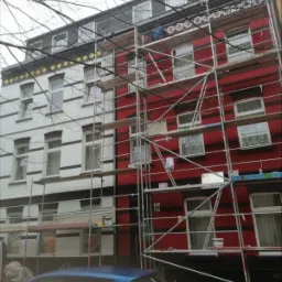 Budynek z rusztowaniem, jedna część biała z czarnymi pasami, druga czerwona z czarnymi pasami, widoczny pracownik na rusztowaniu przy górnych zdobieniach.
