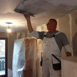 Mężczyzna w białym kombinezonie wyrównuje sufit szpachelką, widoczny otwór na lampę, meble zabezpieczone folią malarską.