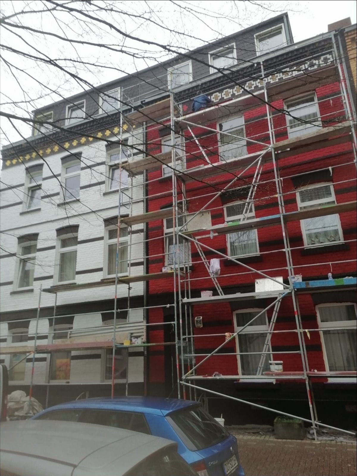 Budynek z rusztowaniem, jedna część biała z czarnymi pasami, druga czerwona z czarnymi pasami, widoczny pracownik na rusztowaniu przy górnych zdobieniach.