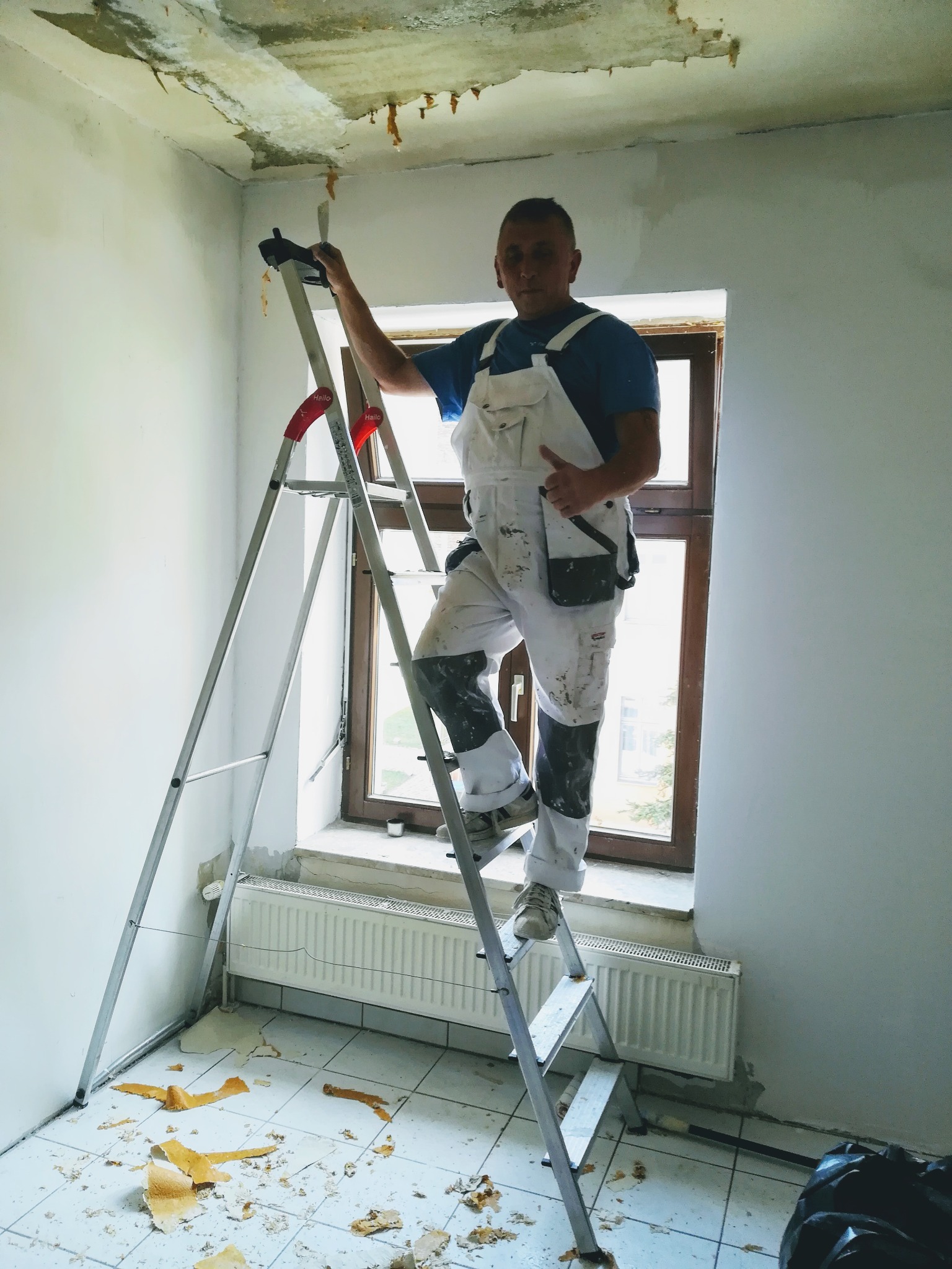 Pracownik w białym kombinezonie stoi na aluminiowej drabinie w trakcie remontu pokoju, widoczne ślady po usuniętej tapecie na suficie i podłodze, okno z widokiem na zewnątrz.