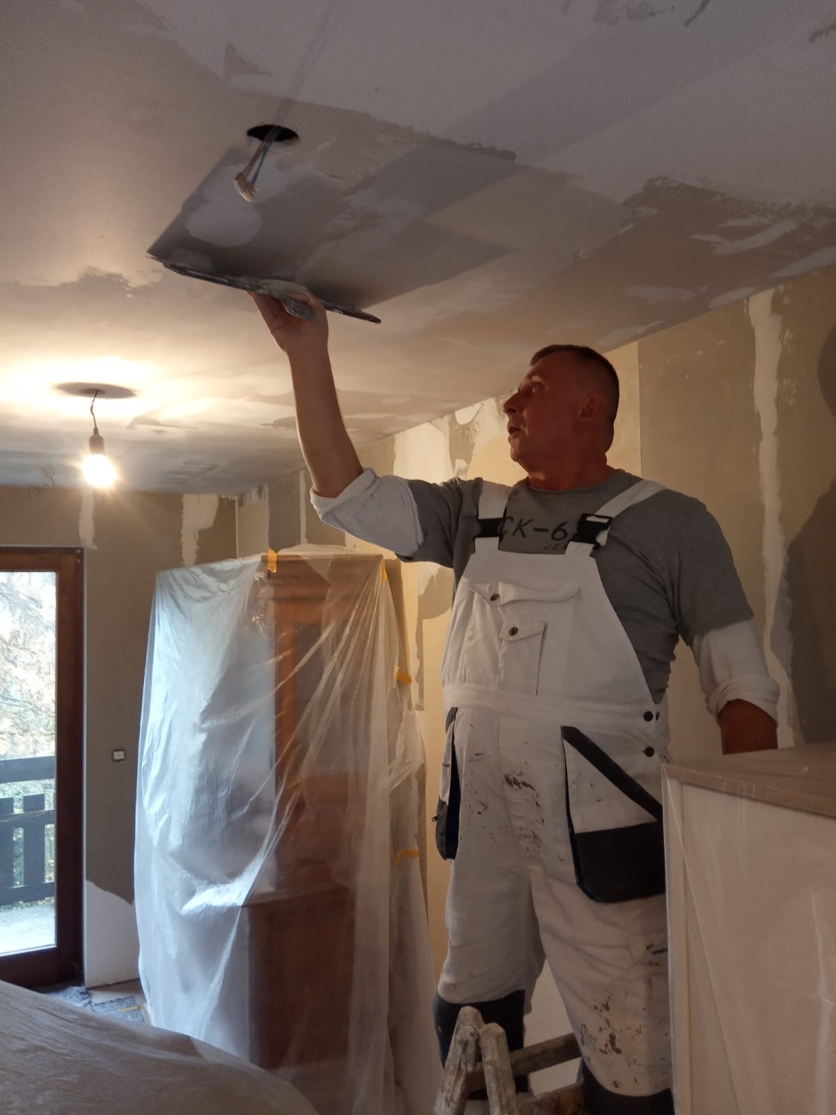 Mężczyzna w białym kombinezonie wyrównuje sufit szpachelką, widoczny otwór na lampę, meble zabezpieczone folią malarską.