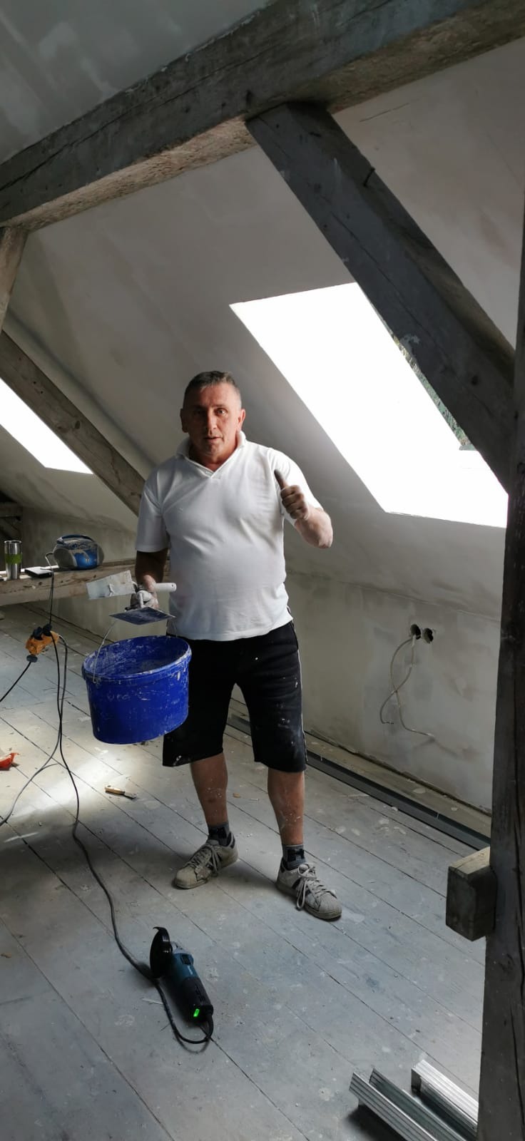 Mężczyzna w białej koszulce i czarnych spodenkach, z uniesionym kciukiem, trzymający niebieskie wiadro i szpachelkę, na tle poddasza z drewnianymi belkami i częściowo wykończonymi ścianami.