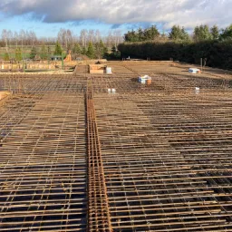 Zbrojenie płyty fundamentowej z widocznymi prętami stalowymi, siatką zbrojeniową i elementami szalunku, w tle zieleń i zabudowa.
