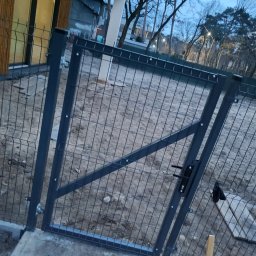 CEZ-BUD. - Nowo zamontowana, ocynkowana furtka z paneli ogrodzeniowych w kolorze grafitowym, widoczna z bliska na tle przygotowanego terenu pod trawnik.