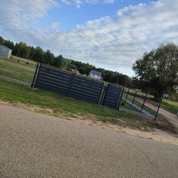 CEZ-BUD. - Nowoczesne, poziome ogrodzenie panelowe w kolorze grafitowym, z bramą wjazdową, na tle zielonego trawnika i błękitnego nieba z chmurami.