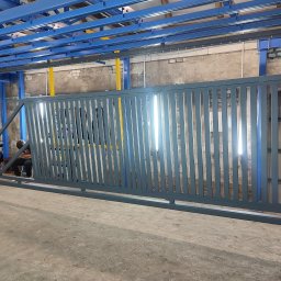 CEZ-BUD. - Szara, przesuwna brama metalowa z pionowymi szczebelkami, widoczna w hali produkcyjnej z niebieską konstrukcją sufitową i fragmentem osoby w tle.