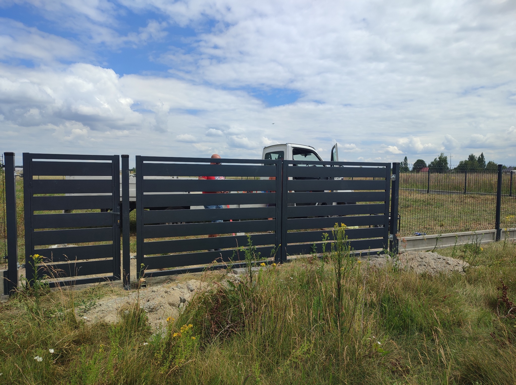 Nowoczesna, pozioma brama wjazdowa i przęsło ogrodzeniowe w kolorze antracytowym, widoczne z zewnątrz posesji, z otwartą furtką i samochodem dostawczym w tle, na tle lekko zachmurzonego nieba.
