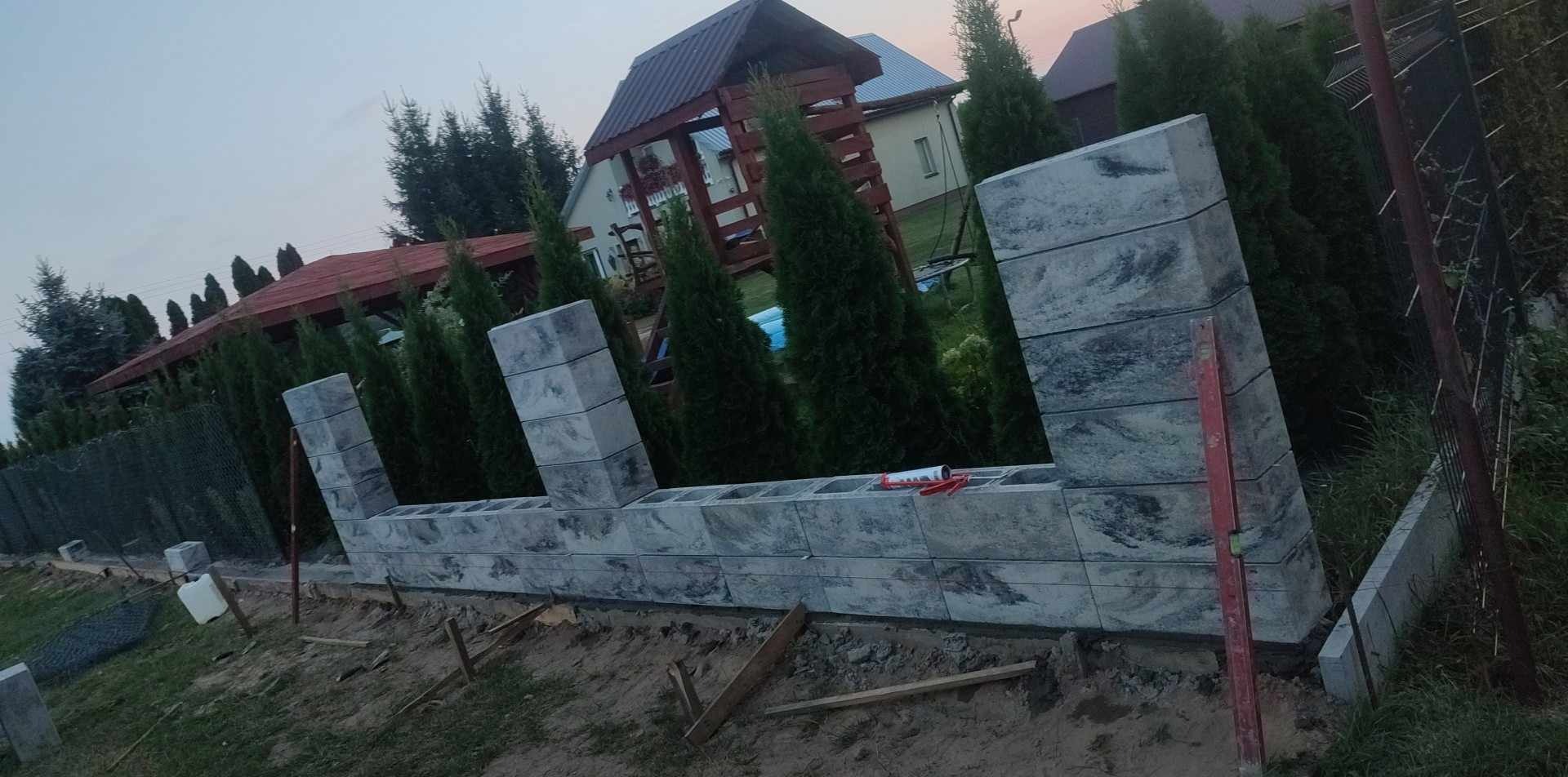 Wznoszenie ogrodzenia z szarych, betonowych bloczków z widocznym klejem montażowym, poziomica i częściowo ułożona podmurówka na tle ogrodu z tujami i altaną.
