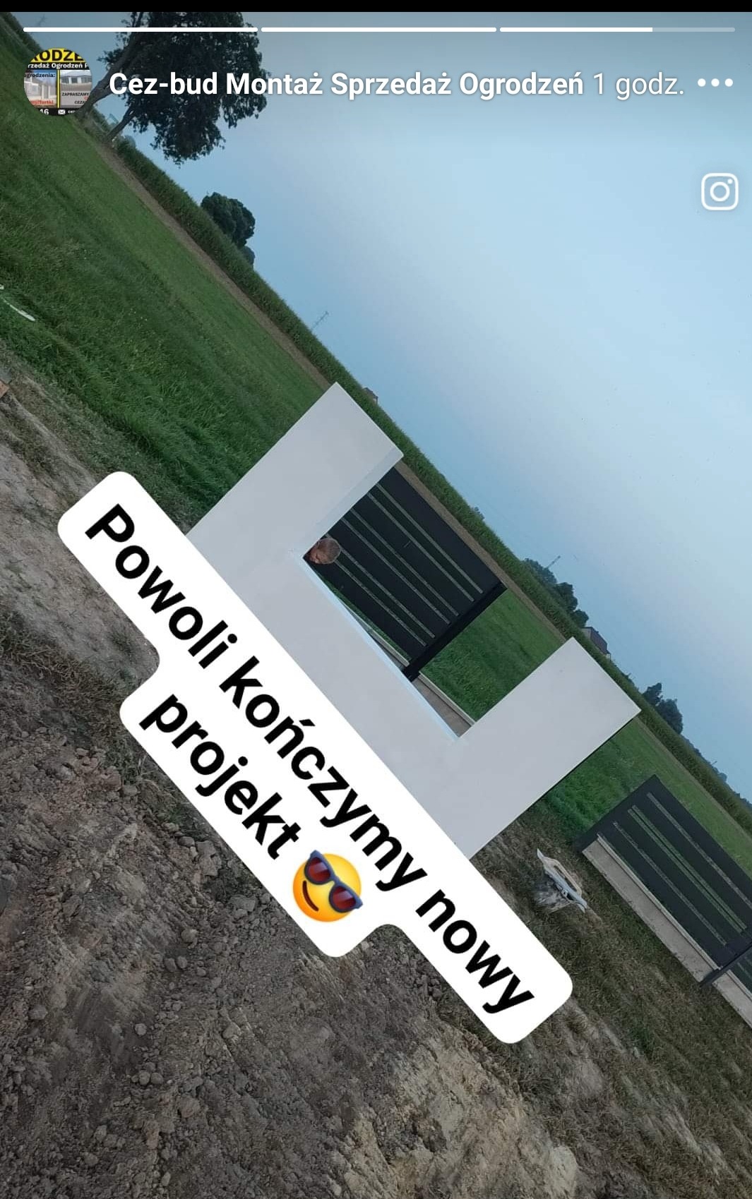 Fragment nowoczesnego, białego ogrodzenia z czarnymi, poziomymi panelami, widziany pod kątem na tle zielonego pola i pochmurnego nieba. W tle widoczna osoba. Na zdjęciu naklejka z tekstem 'Powoli...