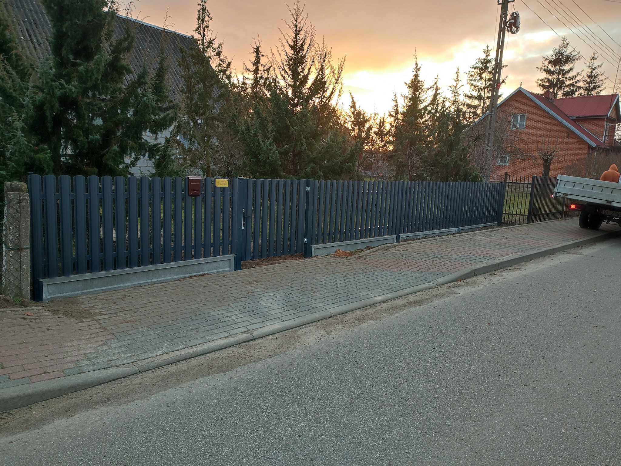Nowoczesne, szare ogrodzenie ze stalowych paneli pionowych, z podmurówką betonową, na tle domu z czerwonej cegły i zachodzącego słońca, widoczne z poziomu ulicy.