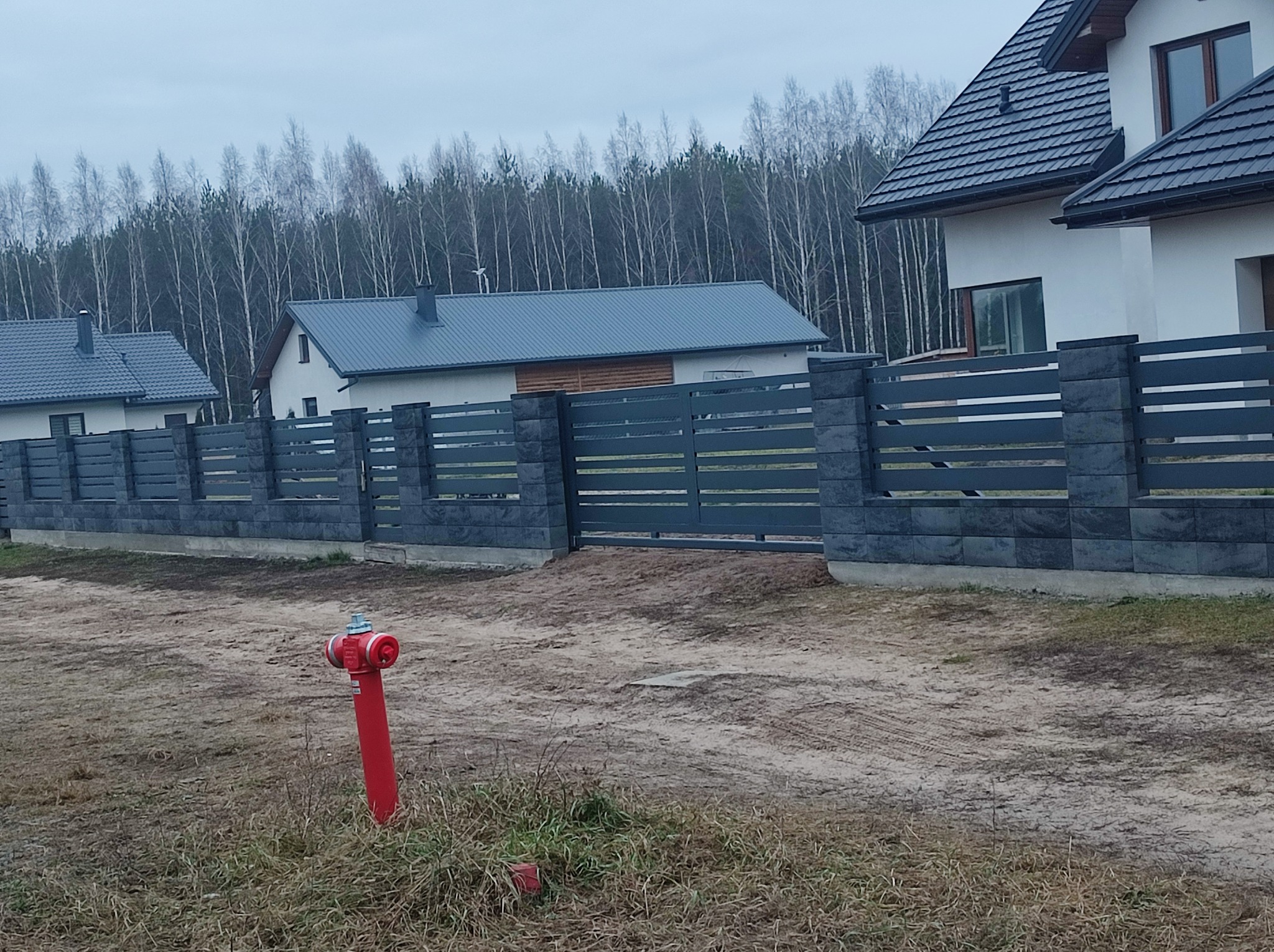 Nowoczesne, poziome, szare ogrodzenie panelowe z bramą przesuwną, widoczne na tle domów i lasu, z hydrantem w pierwszym planie.
