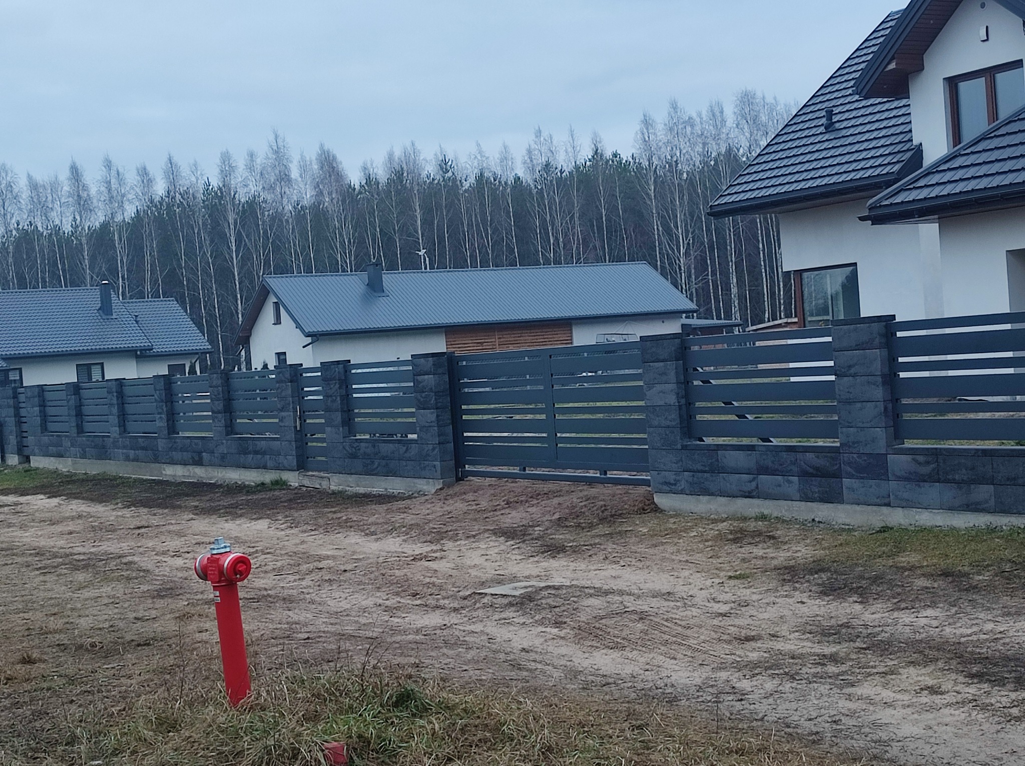Nowoczesne ogrodzenie panelowe z betonowymi słupkami w kolorze grafitowym, widok na domy jednorodzinne i las w tle, czerwony hydrant na pierwszym planie.