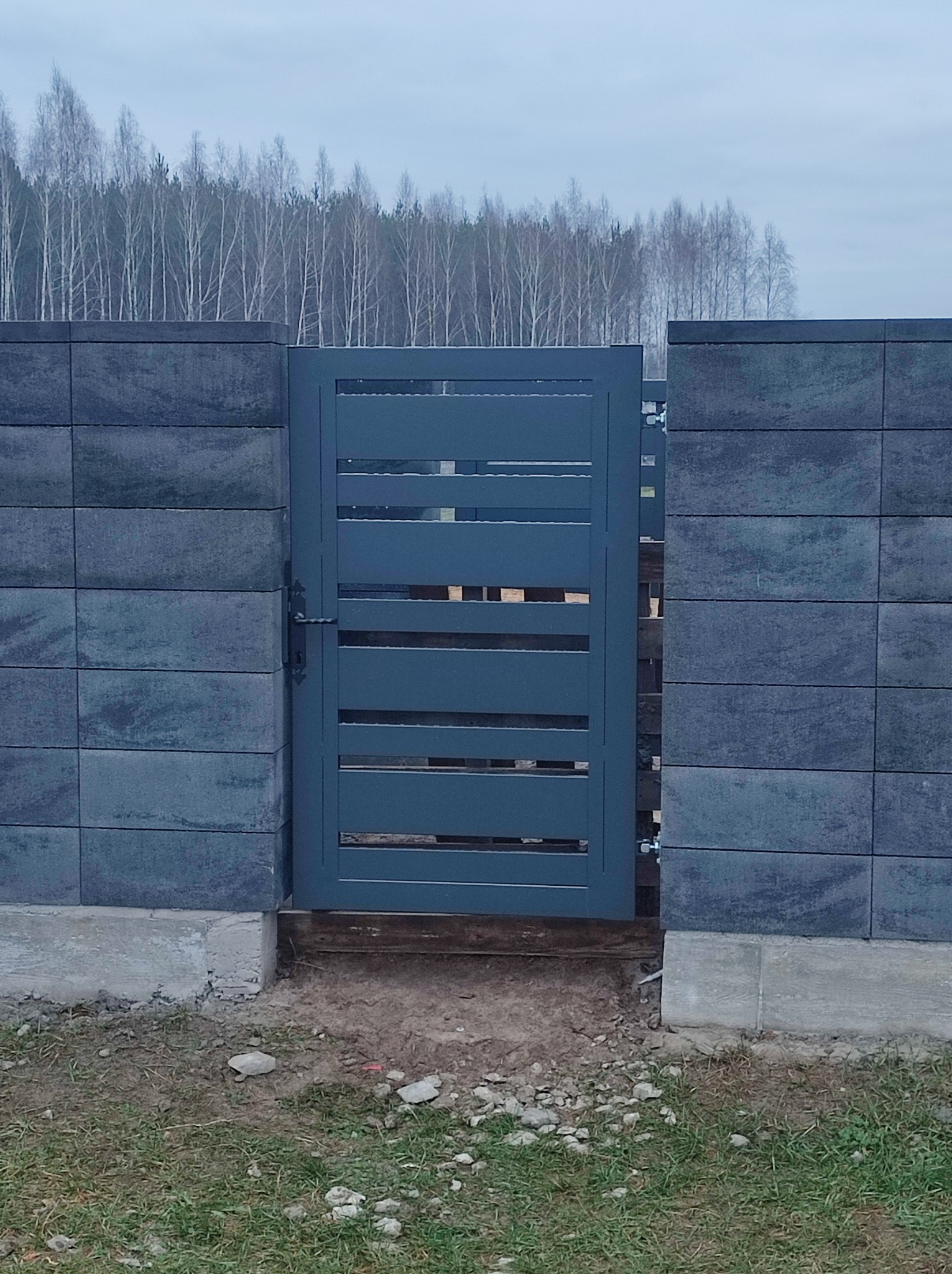 Nowoczesna brama panelowa w kolorze grafitowym, zamontowana w ogrodzeniu z betonowych bloczków, widok na las w tle.