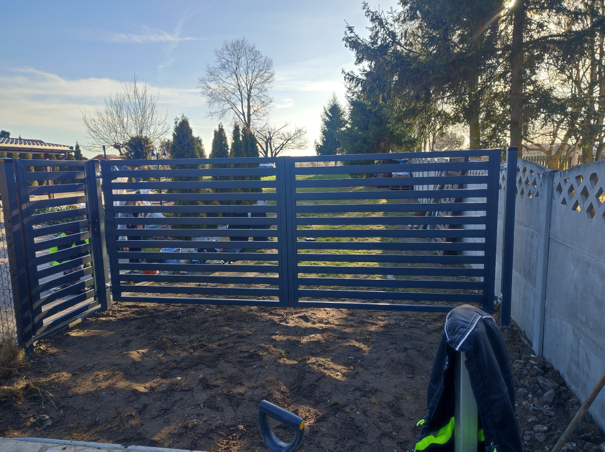 Nowoczesna, pozioma brama panelowa w kolorze antracytowym z otwartą furtką, widok na podwórze z drzewami, obok betonowy płot, na ziemi leżąca kurtka robocza i szpadel.