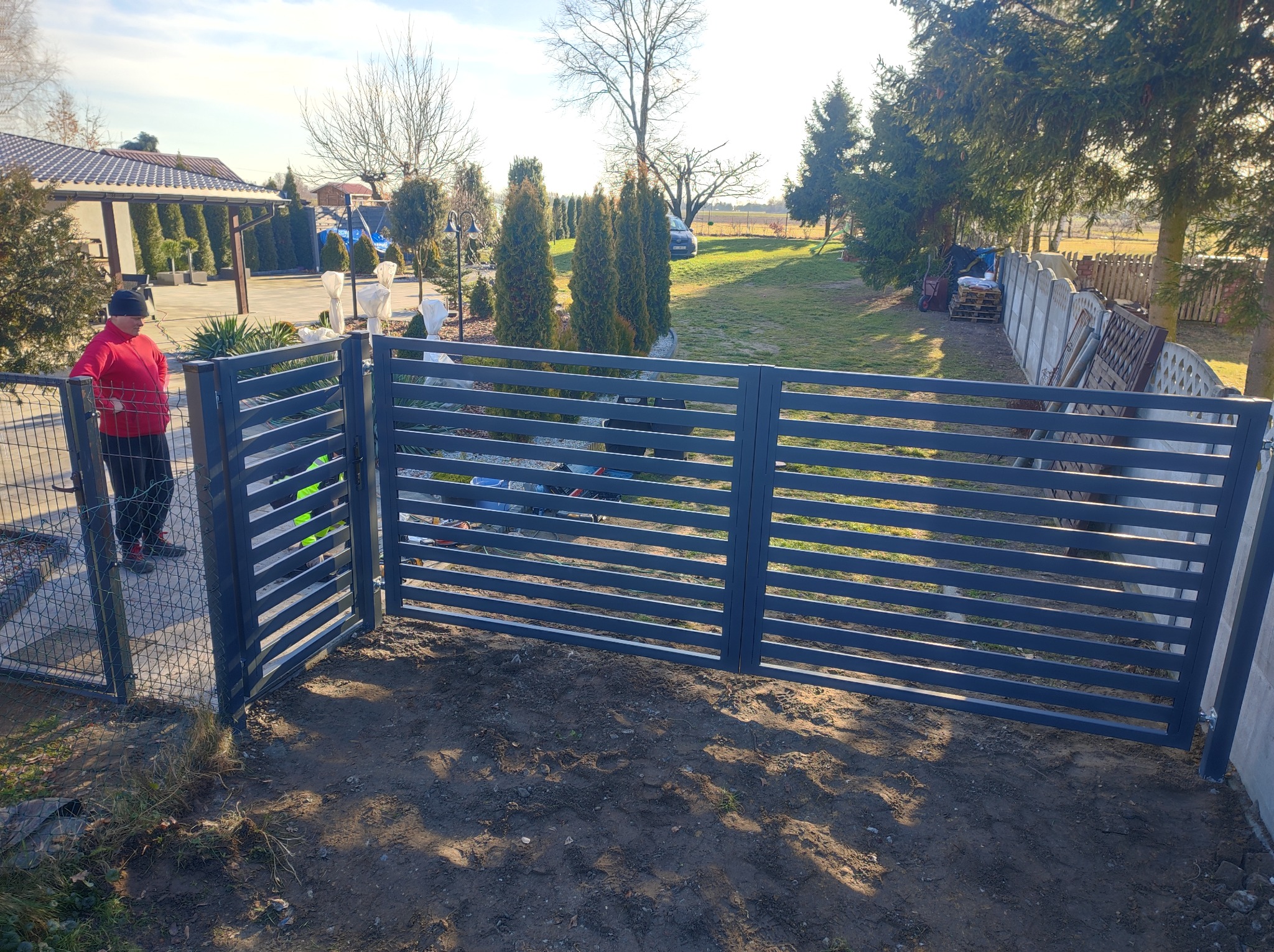 Nowoczesne, szare ogrodzenie panelowe z otwartą furtką wejściową, widok na posesję z tujami i domem w tle, w pobliżu stoi osoba w czerwonej bluzie.
