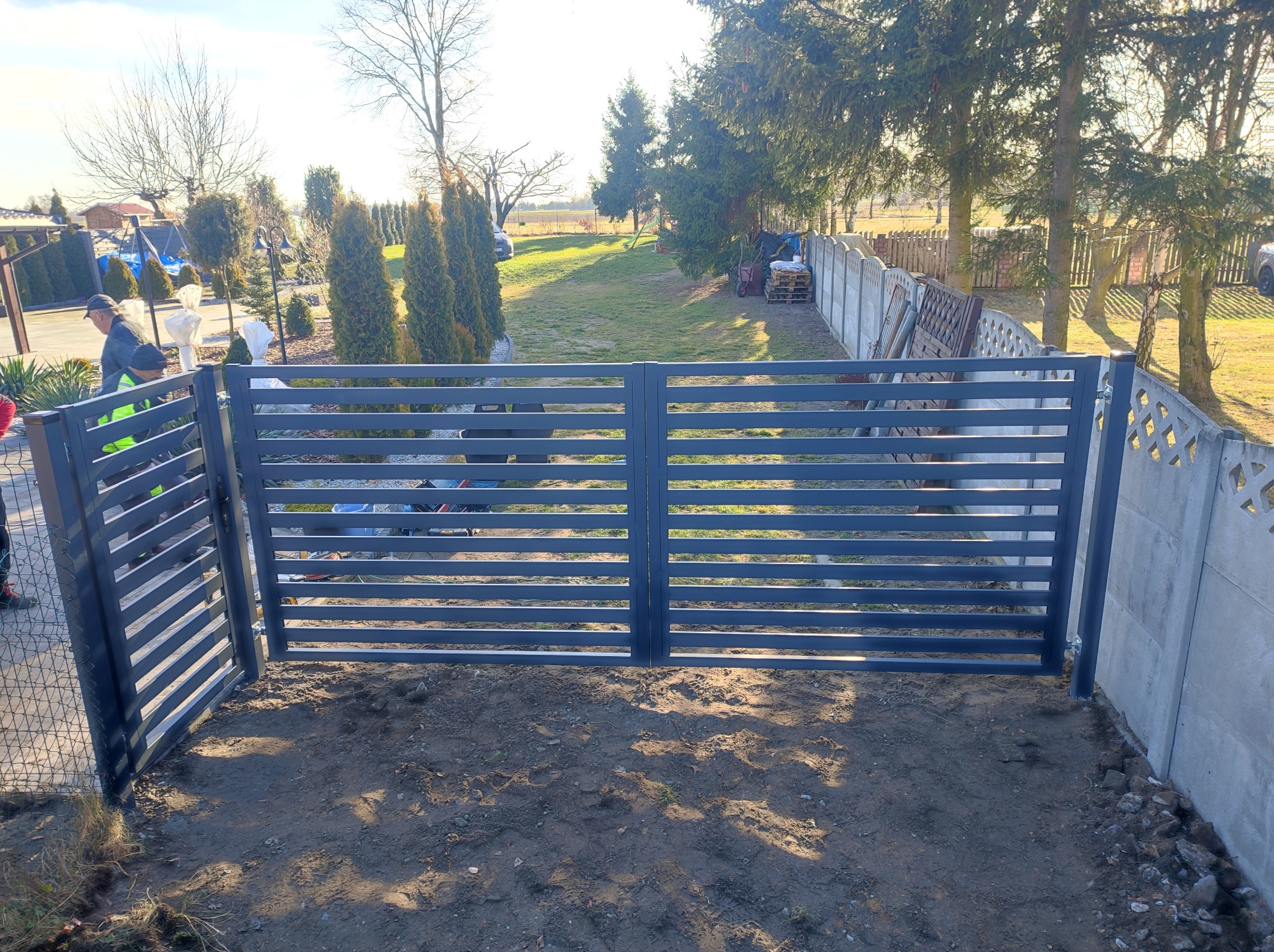 Nowoczesna, grafitowa brama panelowa z poziomymi szczeblami, zamontowana między betonowym a drewnianym ogrodzeniem na nierównym terenie z widocznymi śladami prac montażowych.