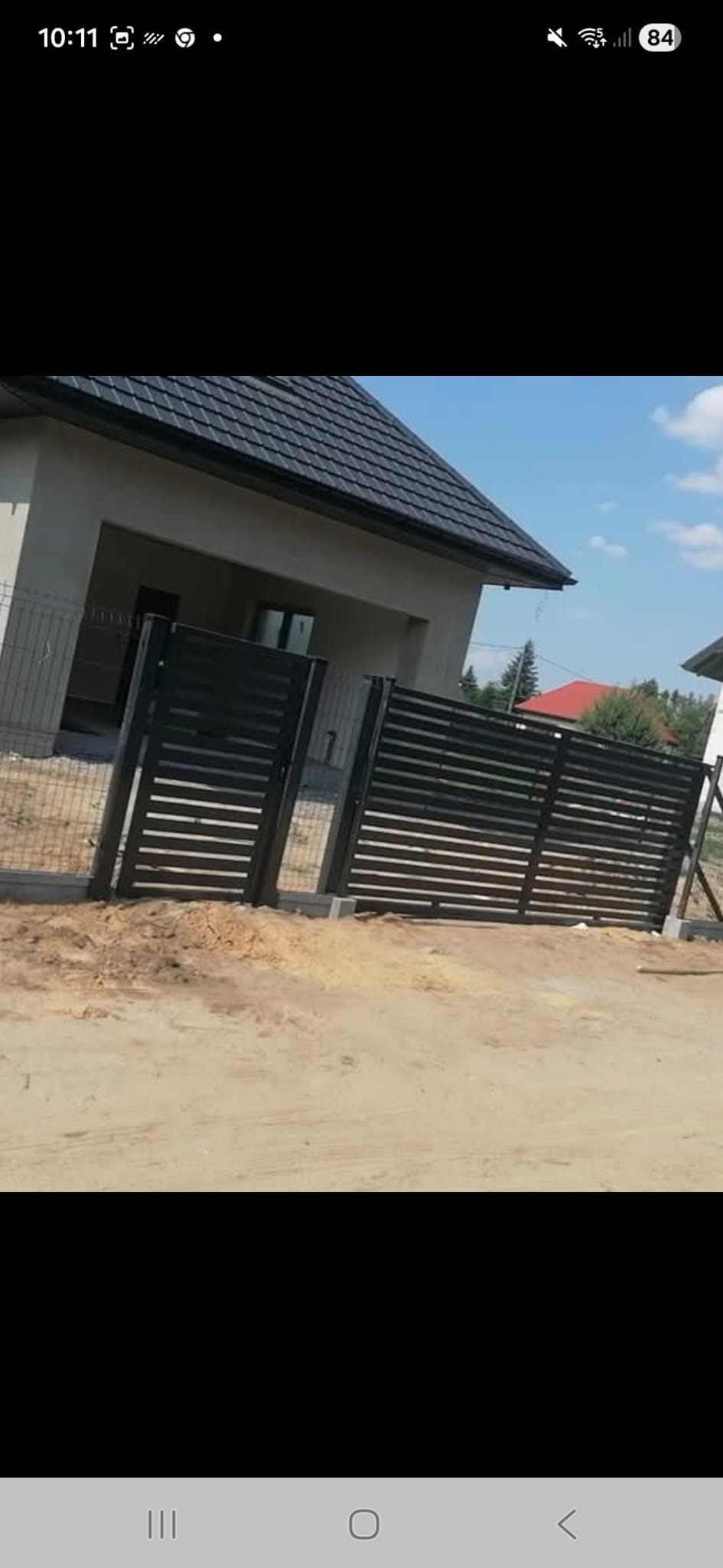 Nowoczesne, poziome ogrodzenie panelowe w kolorze antracytowym przed domem z szarą elewacją i ciemnym dachem. Widoczne słupki i brama wjazdowa.