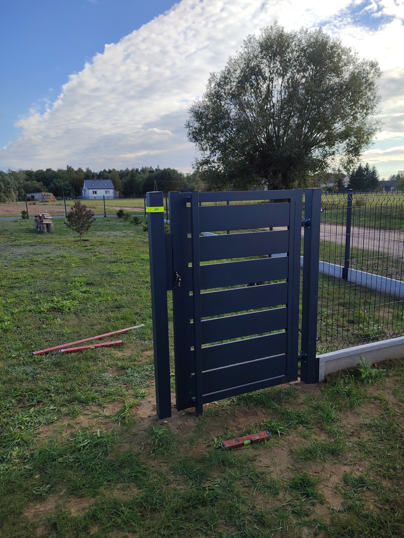 Nowoczesna, grafitowa furtka z poziomymi panelami, zamontowana przy ogrodzeniu panelowym. Na trawie leżą poziomice, w tle dom i drzewa.